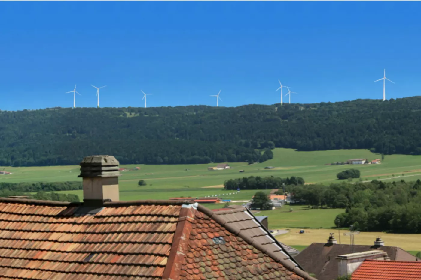 Dachlandschaft mit roten Ziegeln und Windkraftanlagen im Hintergrund, blauer Himmel.