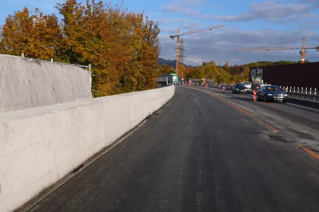Bauarbeiten an einer Straße mit einer Betonwand und herbstlichen Bäumen im Hintergrund.
