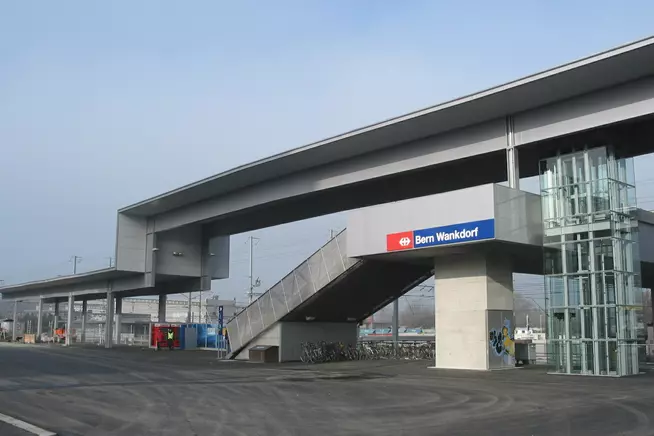 Moderne Haltestelle mit Glasaufzug und überdachten Warteschlangen, bewölkter Himmel.