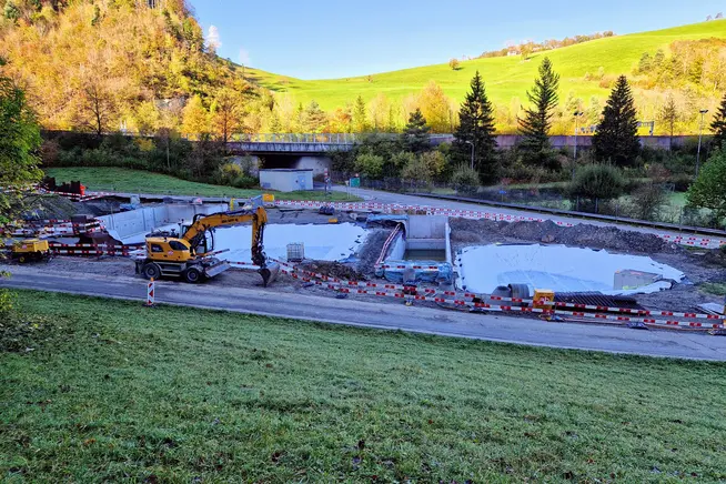 Bauarbeiten an einem Grundstück mit Bagger und Wasserflächen, umgeben von Bäumen.