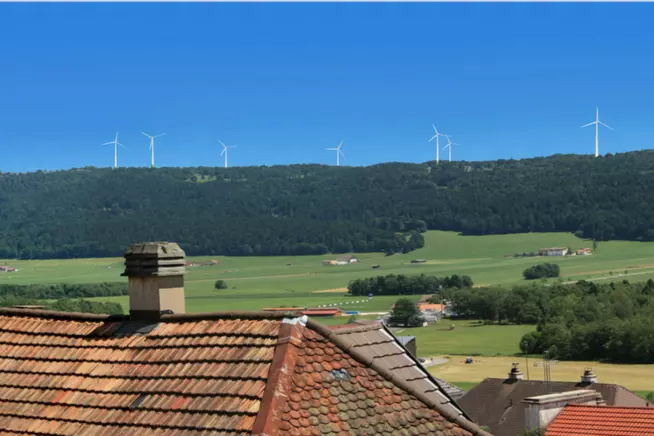 Dachlandschaft mit roten Ziegeln und Windkraftanlagen im Hintergrund, blauer Himmel.