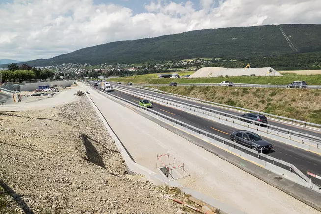 Blick auf eine gut ausgebaute Strasse mit vorbeifahrenden Autos und grünem Hügel im Hintergrund.